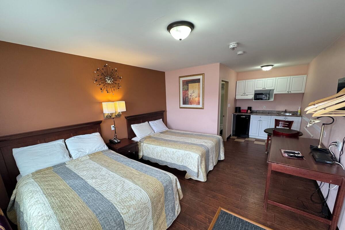 A hotel room with two beds, a desk, a chair, a small kitchenette, and a bathroom visible through an open door.