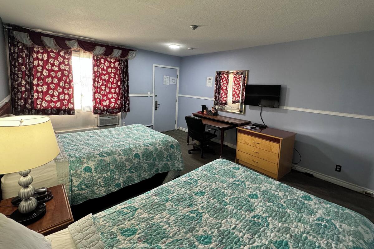 A hotel room with two beds, a desk, a dresser, a TV, and a window with curtains.