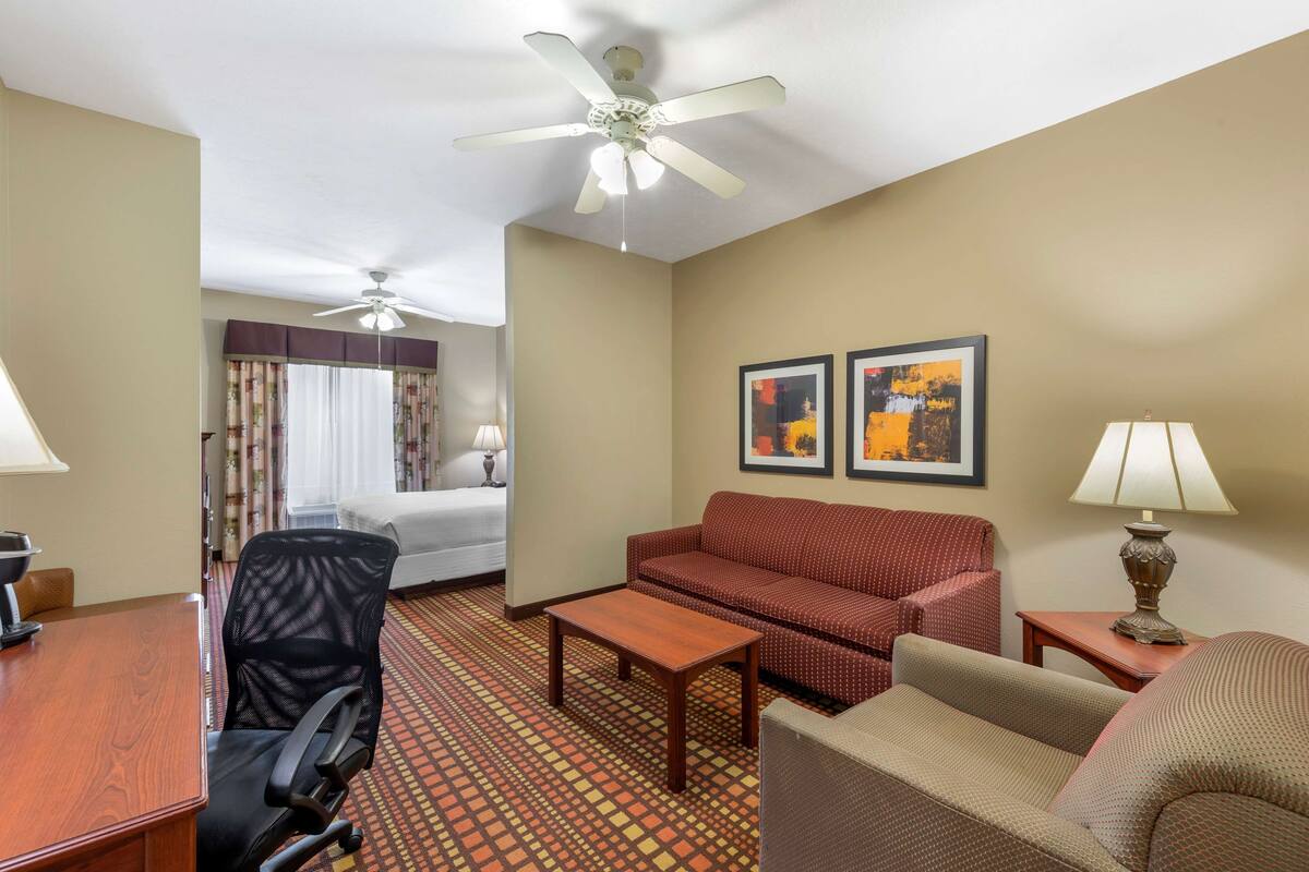 A hotel room with a bed, a desk, a chair, a sofa, a coffee table, a lamp, and two ceiling fans.