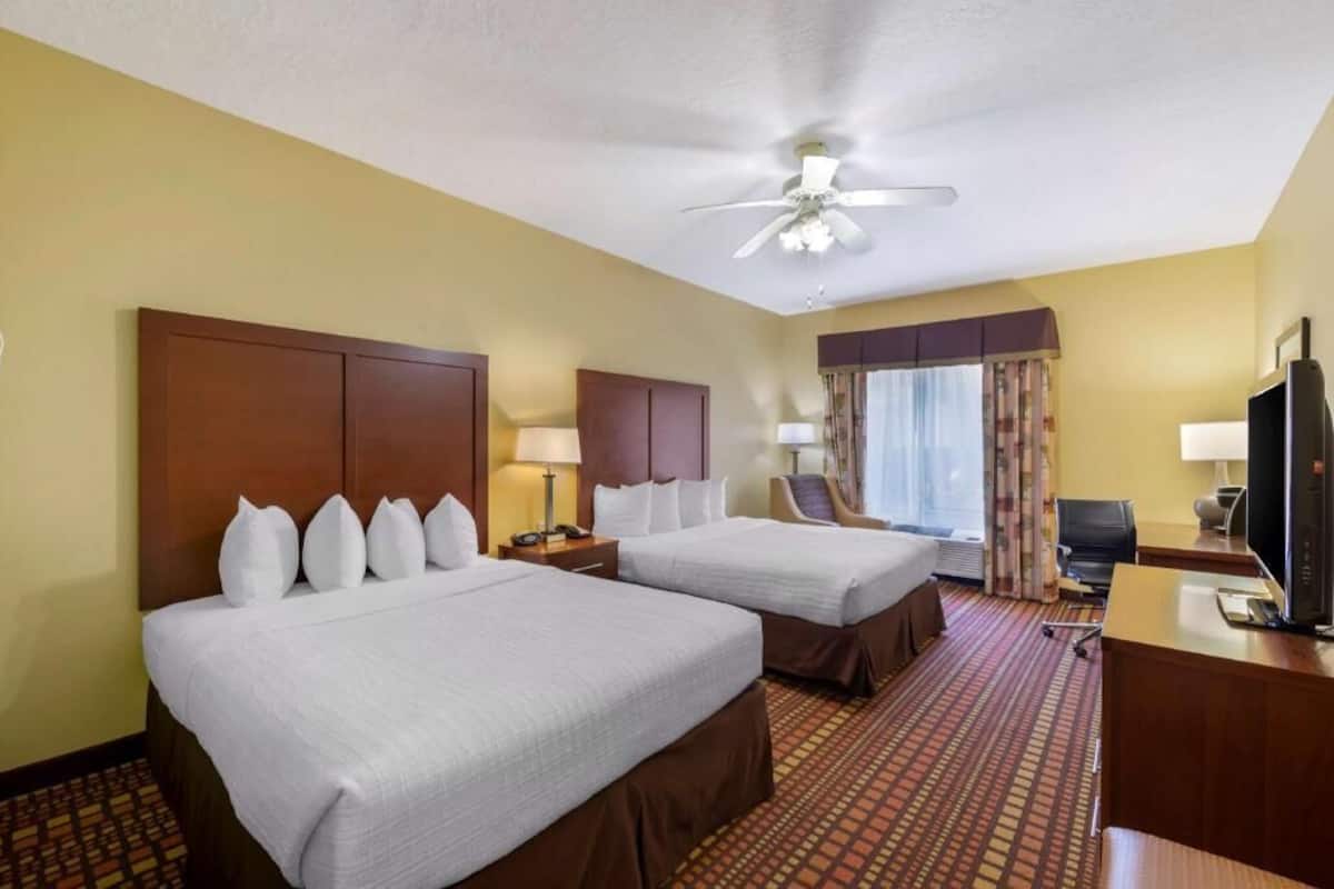 A hotel room with two beds, a desk, a TV, a ceiling fan, and a window with curtains.