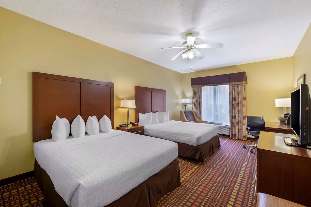 A hotel room with two beds, a desk, a television, a ceiling fan, and a window with curtains.