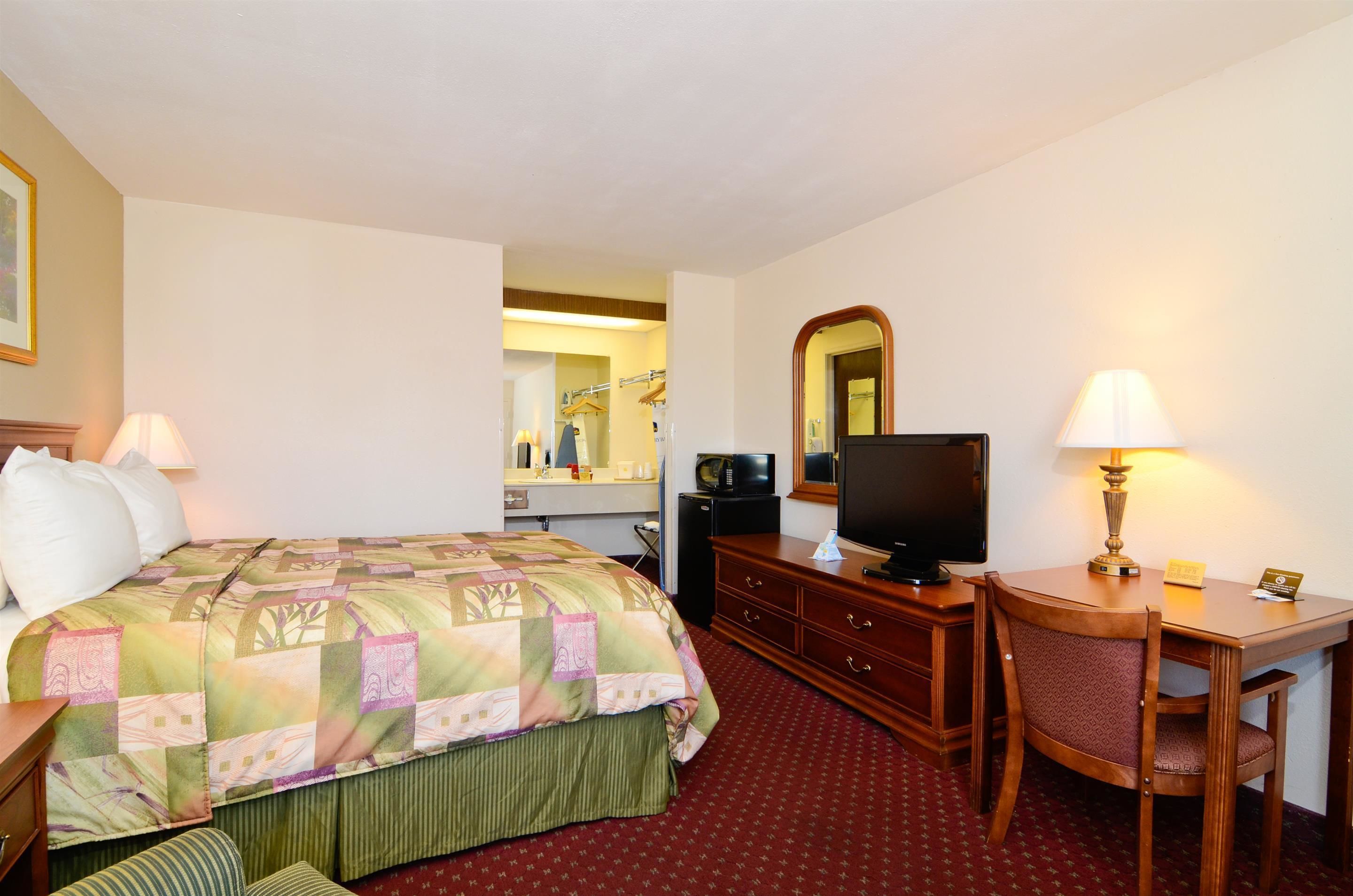 A hotel room with a bed, a desk, a chair, a television, a mirror, and a lamp.