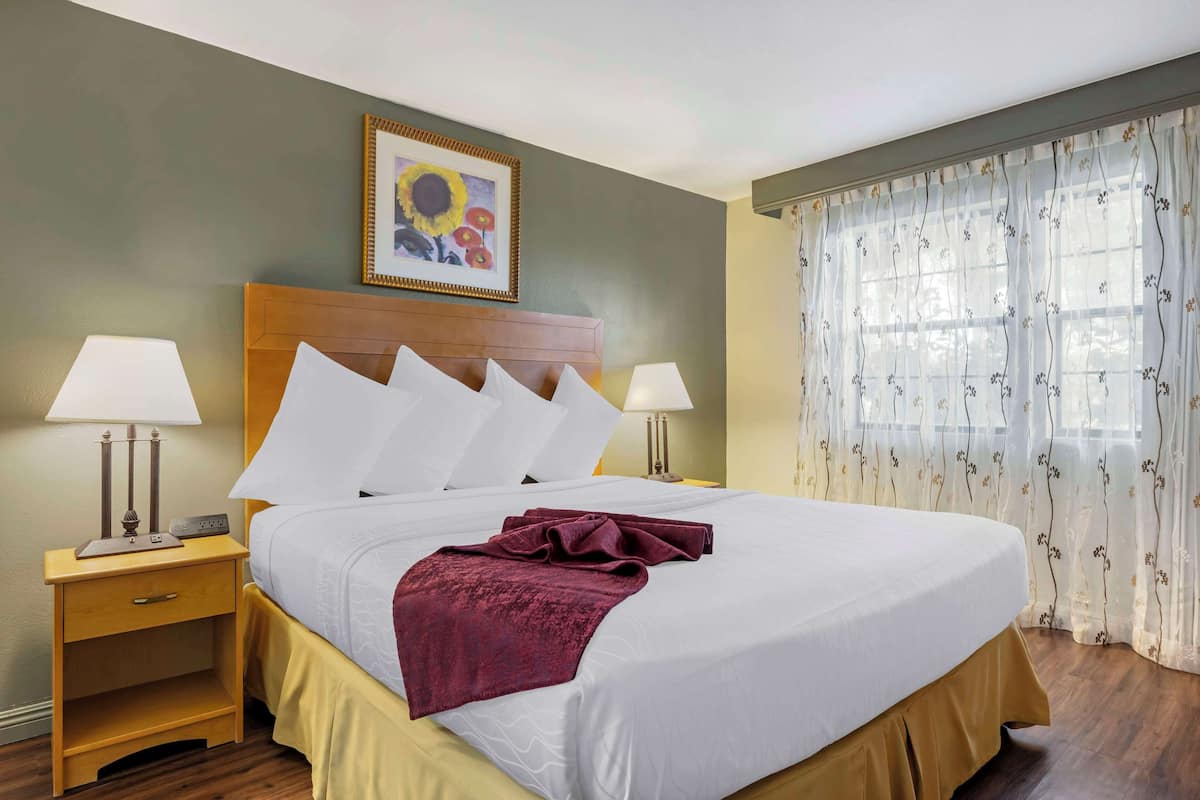 A neatly made bed with white linens and a maroon throw, flanked by two bedside tables with lamps, in a room with a wooden headboard and a framed painting above.
