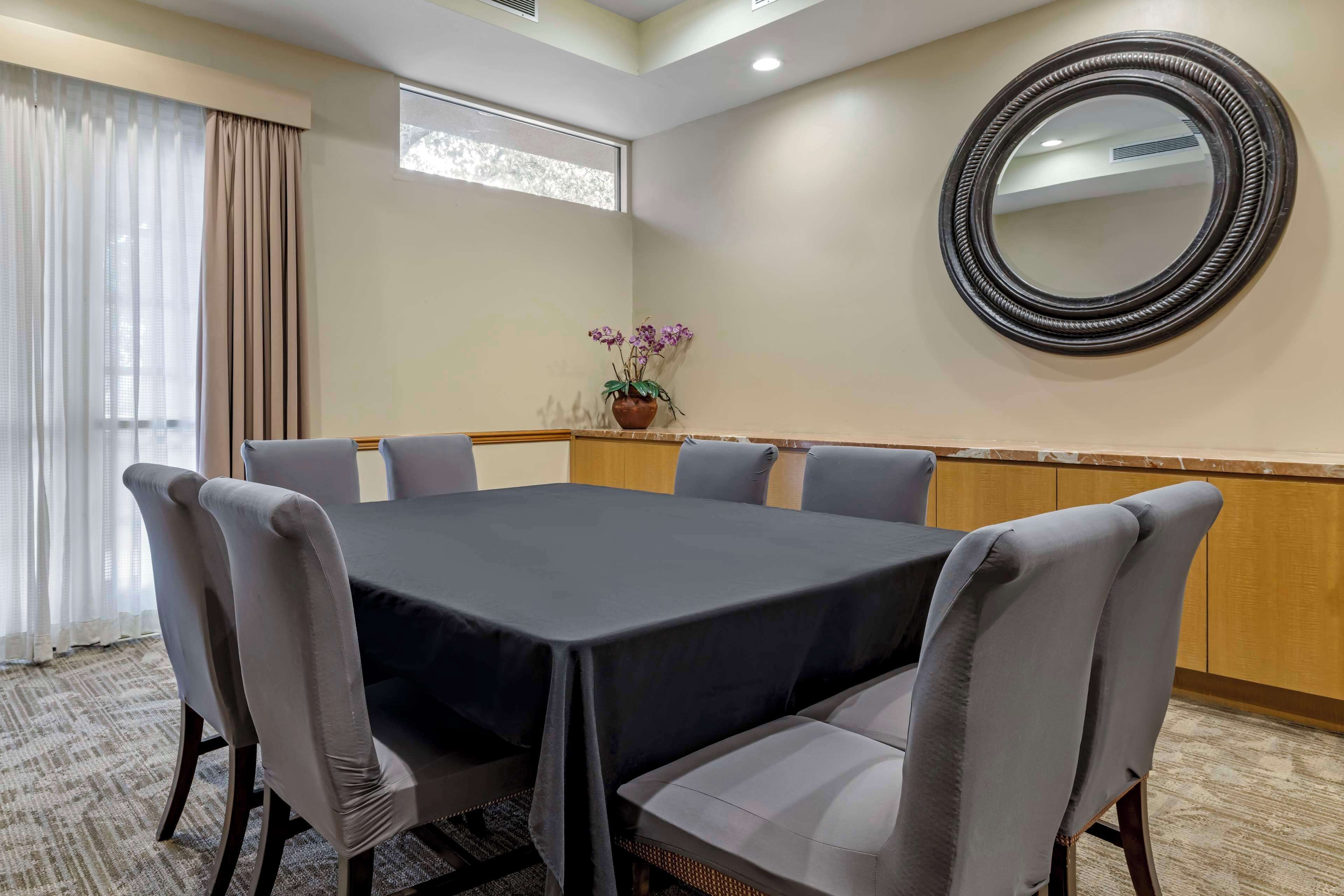 A conference room with a large table, six chairs, a potted plant, a round mirror, and a window with curtains.