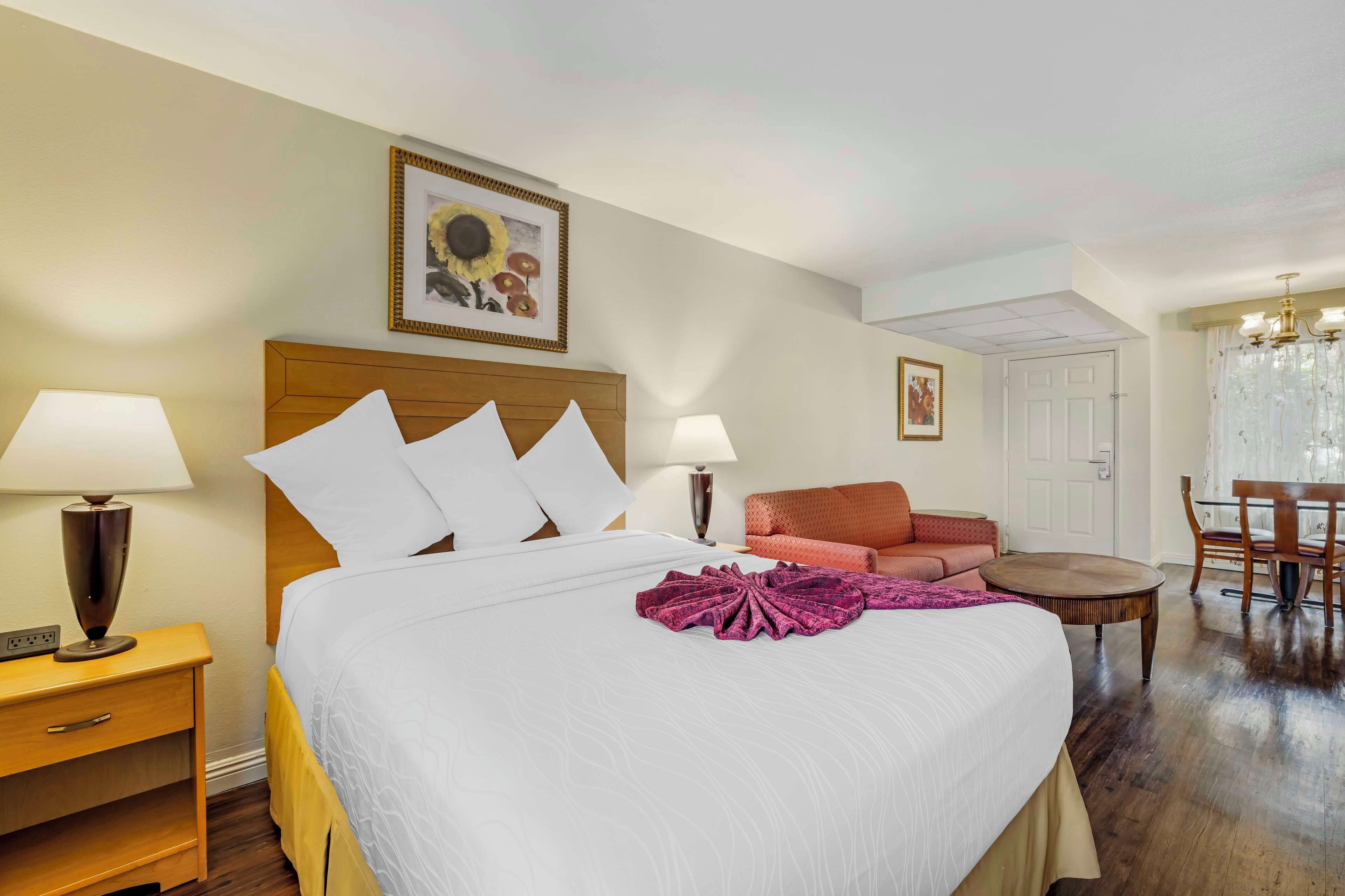 A hotel room with a large bed, two bedside lamps, a wooden nightstand, a red sofa, a round wooden table, and a chandelier.