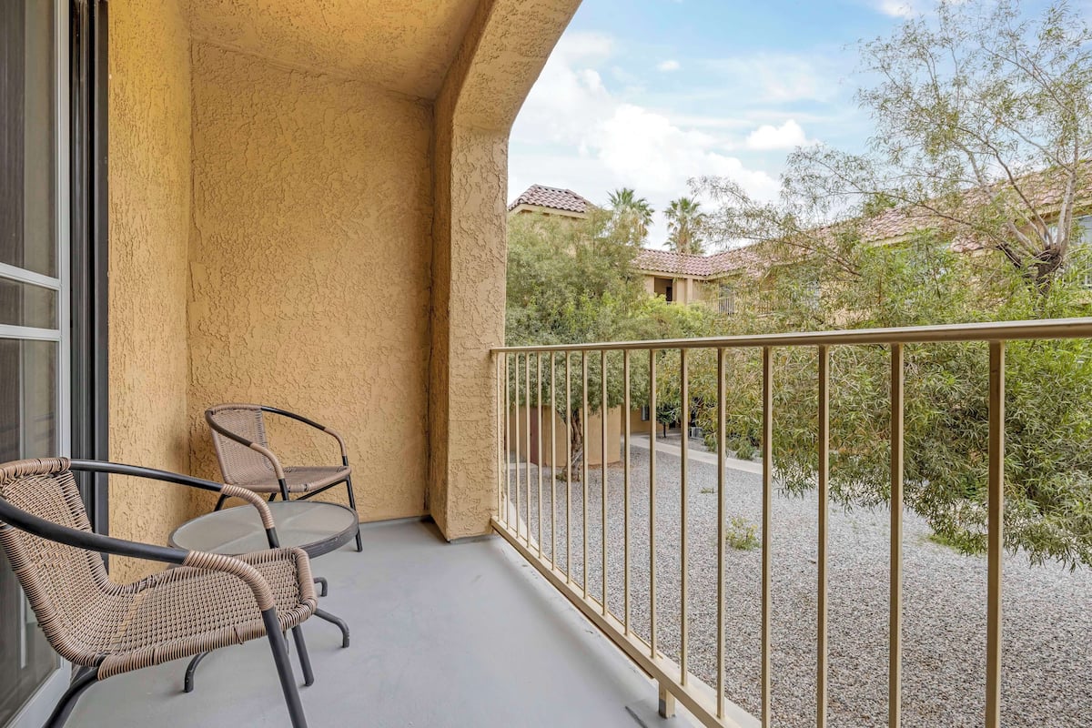 A balcony with wicker chairs and a glass table, overlooking a gravel area with trees and buildings.
