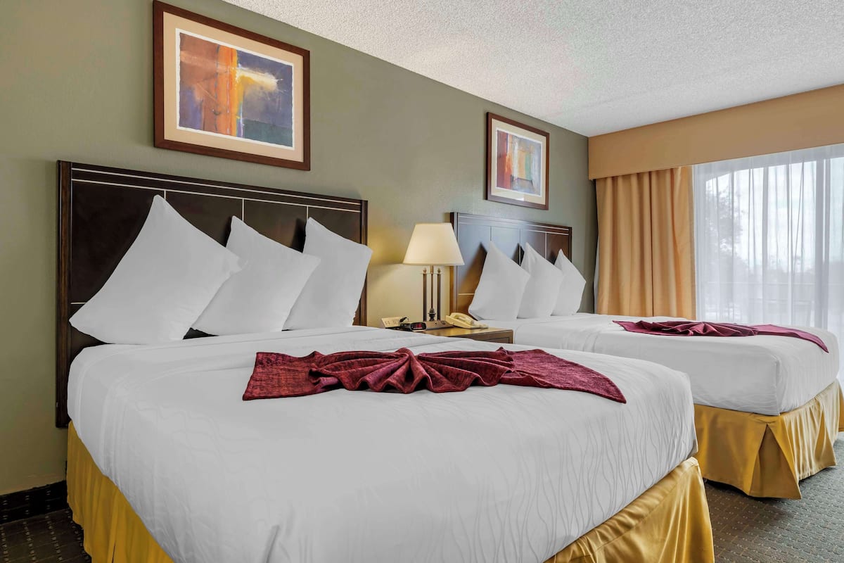 A hotel room with two beds, a nightstand with a lamp, framed artwork on the walls, and a window with curtains.