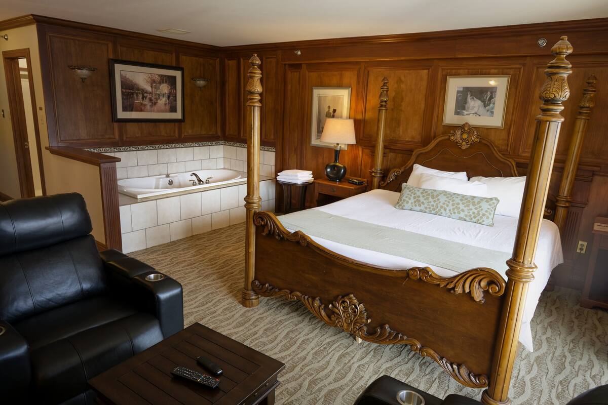 Una habitación de hotel con cama con dosel, una zona de estar con sofá y sillones, una mesita, un baño con lavamanos y bañera, y suelo alfombrado.