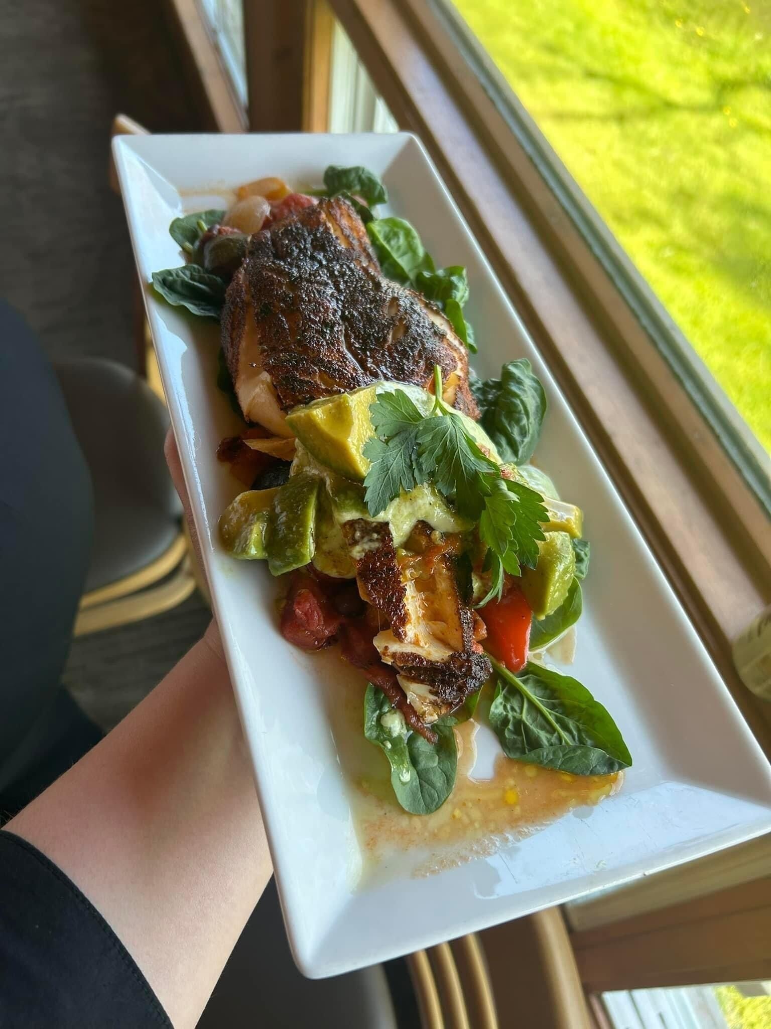 A white rectangular plate with a grilled chicken breast, avocado slices, and greens, held by a person.
