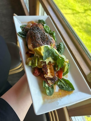 A white rectangular plate with a grilled chicken breast, avocado slices, and greens, held by a person. - Liscombe Lodge Resort and Conference Centre (Liscomb Mills)