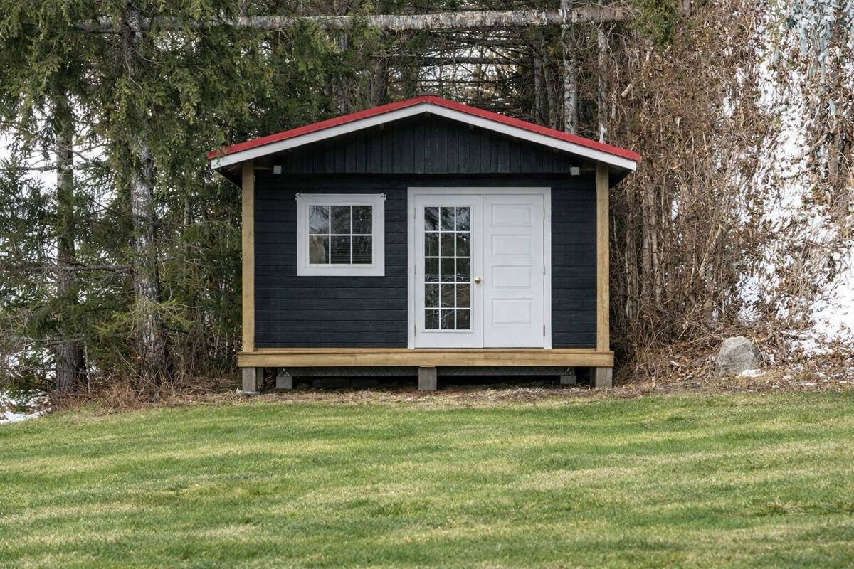 Una pequeña cabaña de madera oscura con techo rojo, puertas y ventanas blancas, rodeada de césped y árboles.