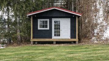 Una pequeña cabaña de madera oscura con techo rojo, puertas y ventanas blancas, rodeada de césped y árboles.