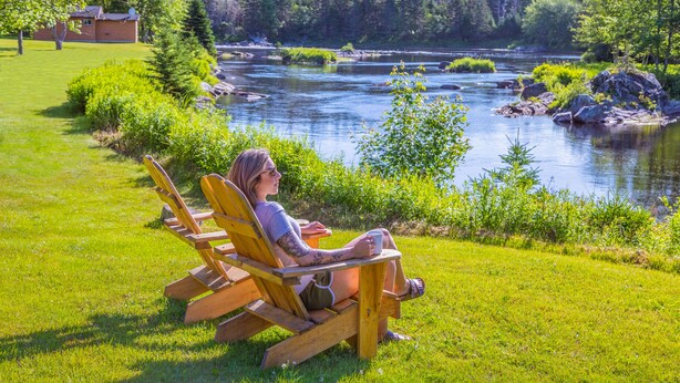 A person sitting on a wooden chair by a river, with a house and forest in the background. - Liscombe Lodge Resort and Conference Centre (Liscomb Mills)