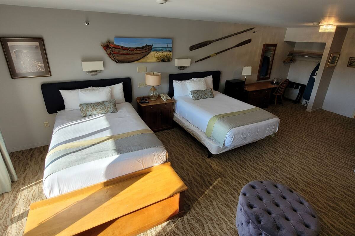 Habitación de hotel con dos camas, un banco de madera, una mesita con lámpara, un cuadro de un barco y un comedor con una silla y una mesa.