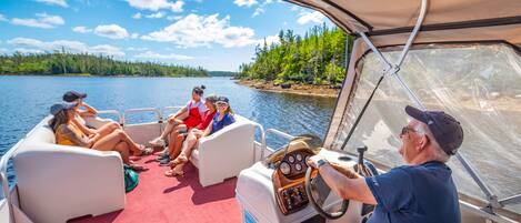 A boat interior with a steering wheel, two cushioned seating areas, and a view of a lake and forest.