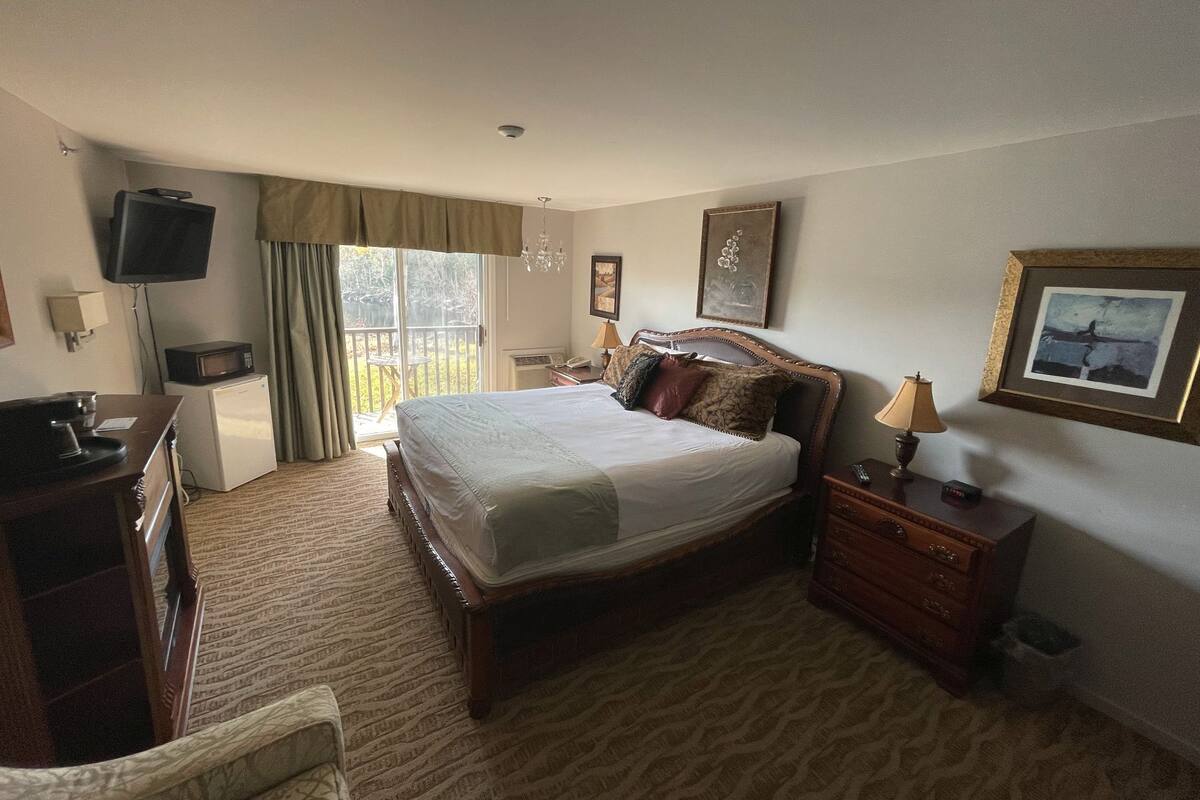 Habitación de hotel con cama, escritorio, televisor, microondas, refrigerador pequeño, silla, mesita de noche con lámpara y un cuadro en la pared.