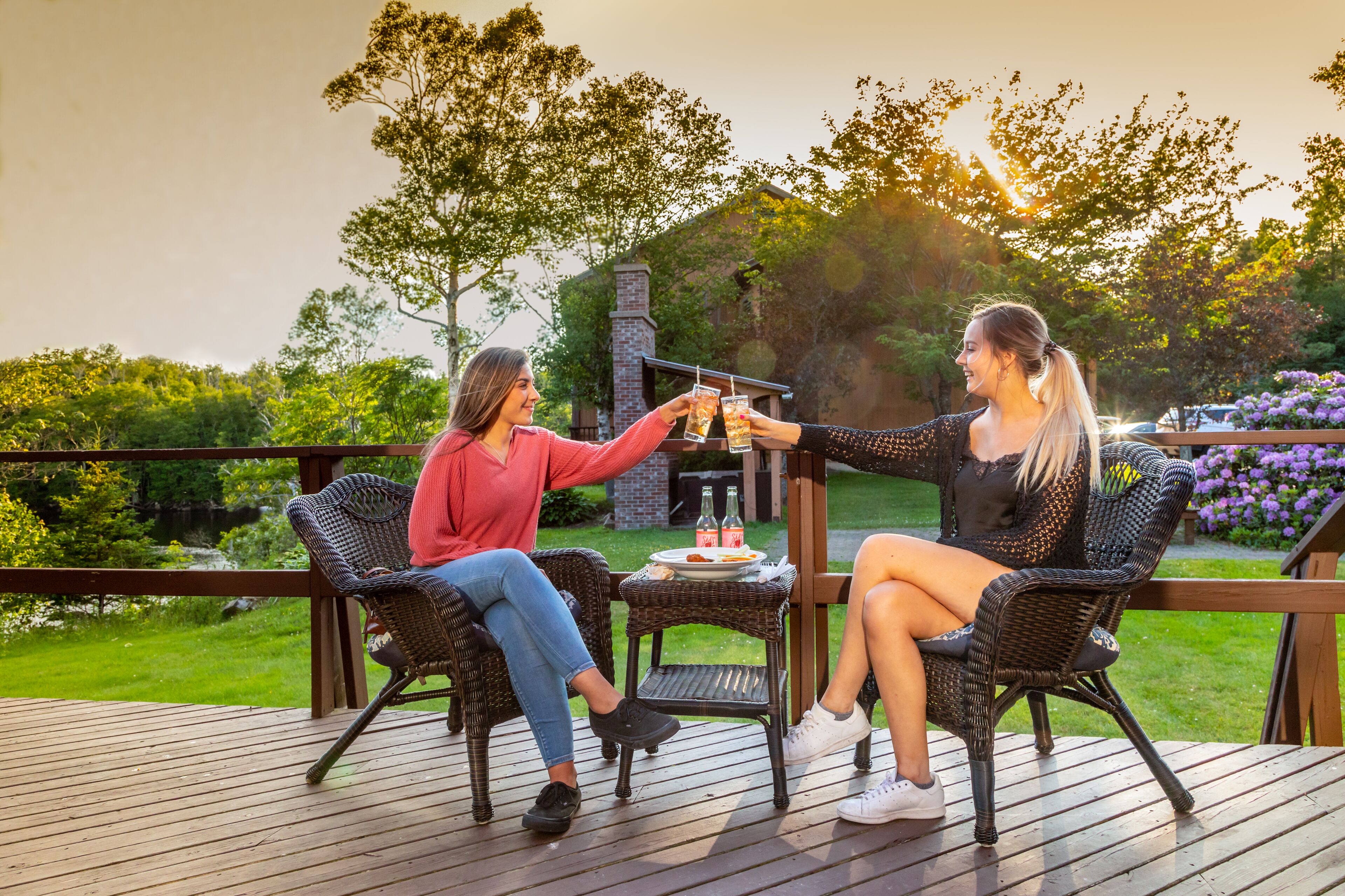 Two people toasting on a wooden deck with wicker chairs, a small table with drinks, and a garden view.