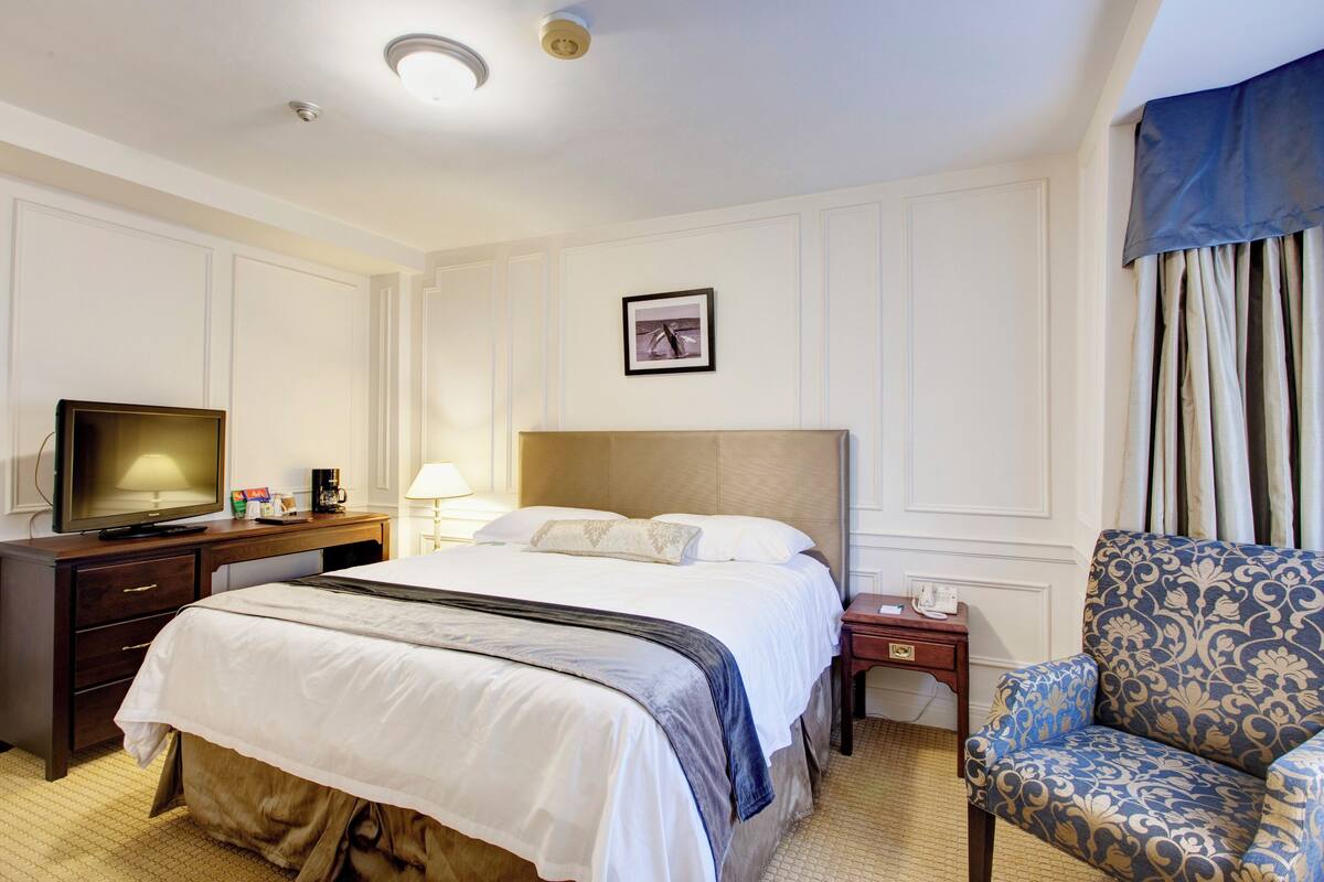 A hotel room with a large bed, a television, a desk with a lamp, and a patterned armchair.
