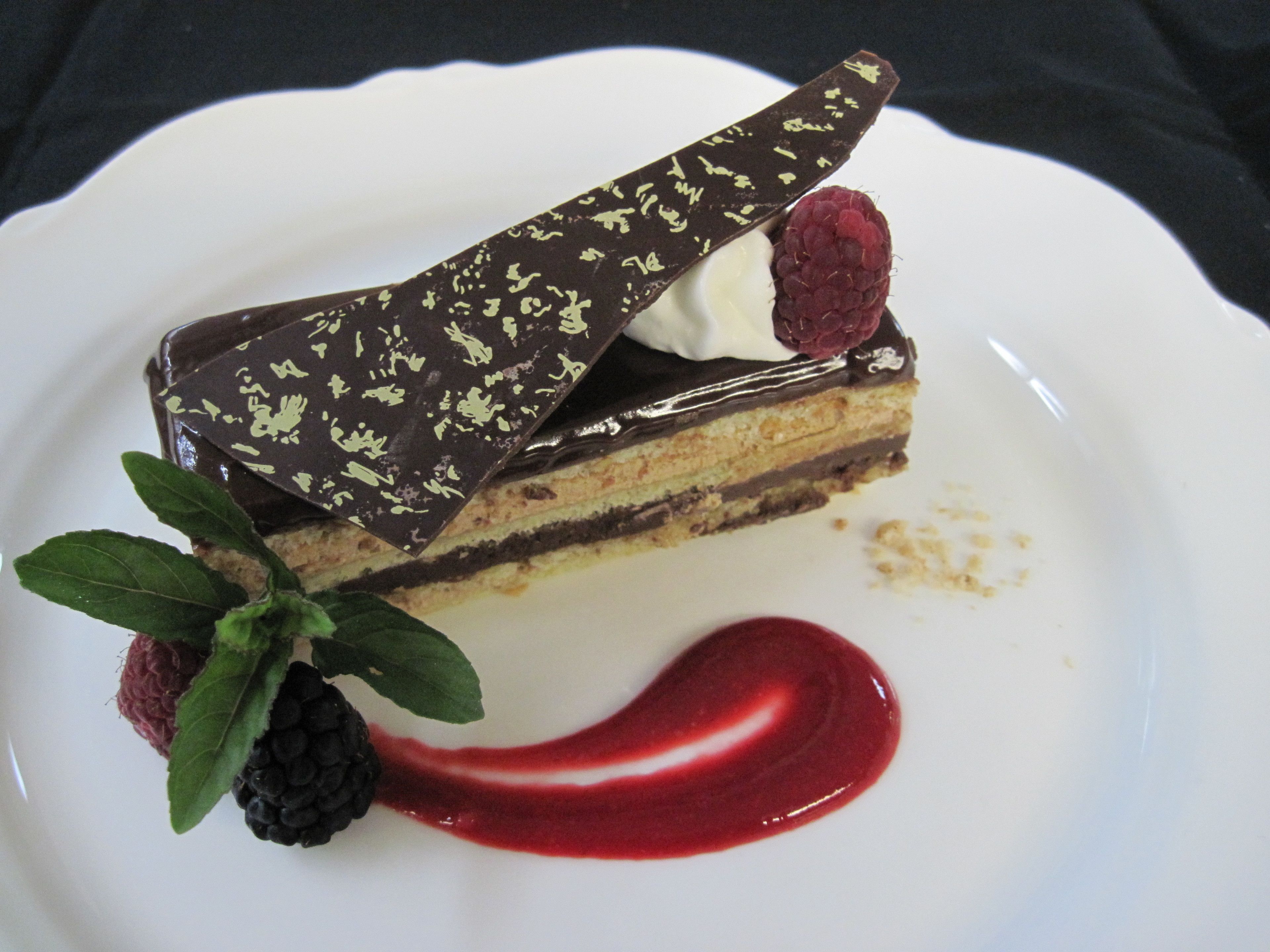 A slice of layered cake with chocolate and raspberry garnish on a white plate.