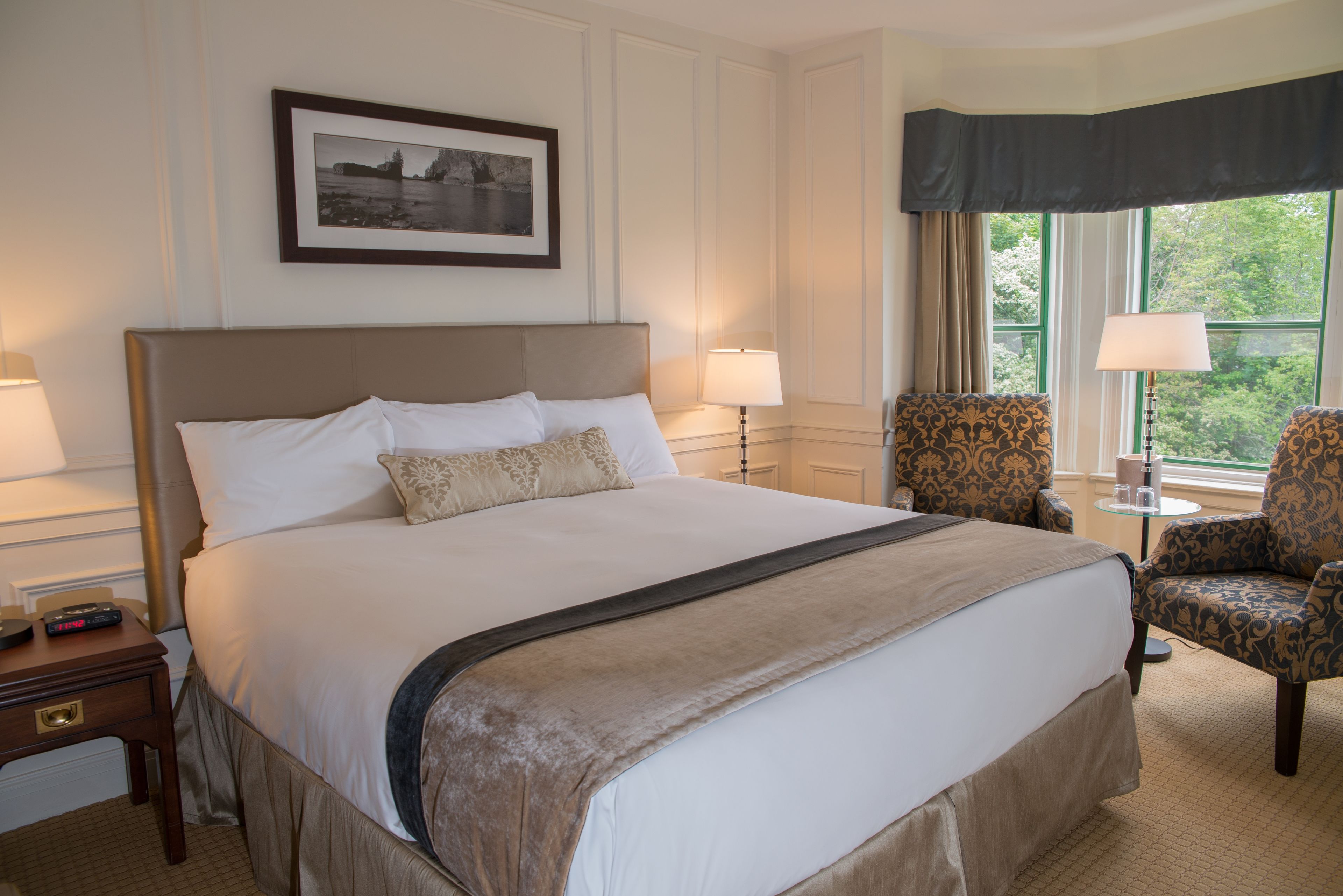 Quarto de hotel com uma cama grande, duas luminárias de cabeceira, uma cadeira, uma mesinha e um quadro na parede.