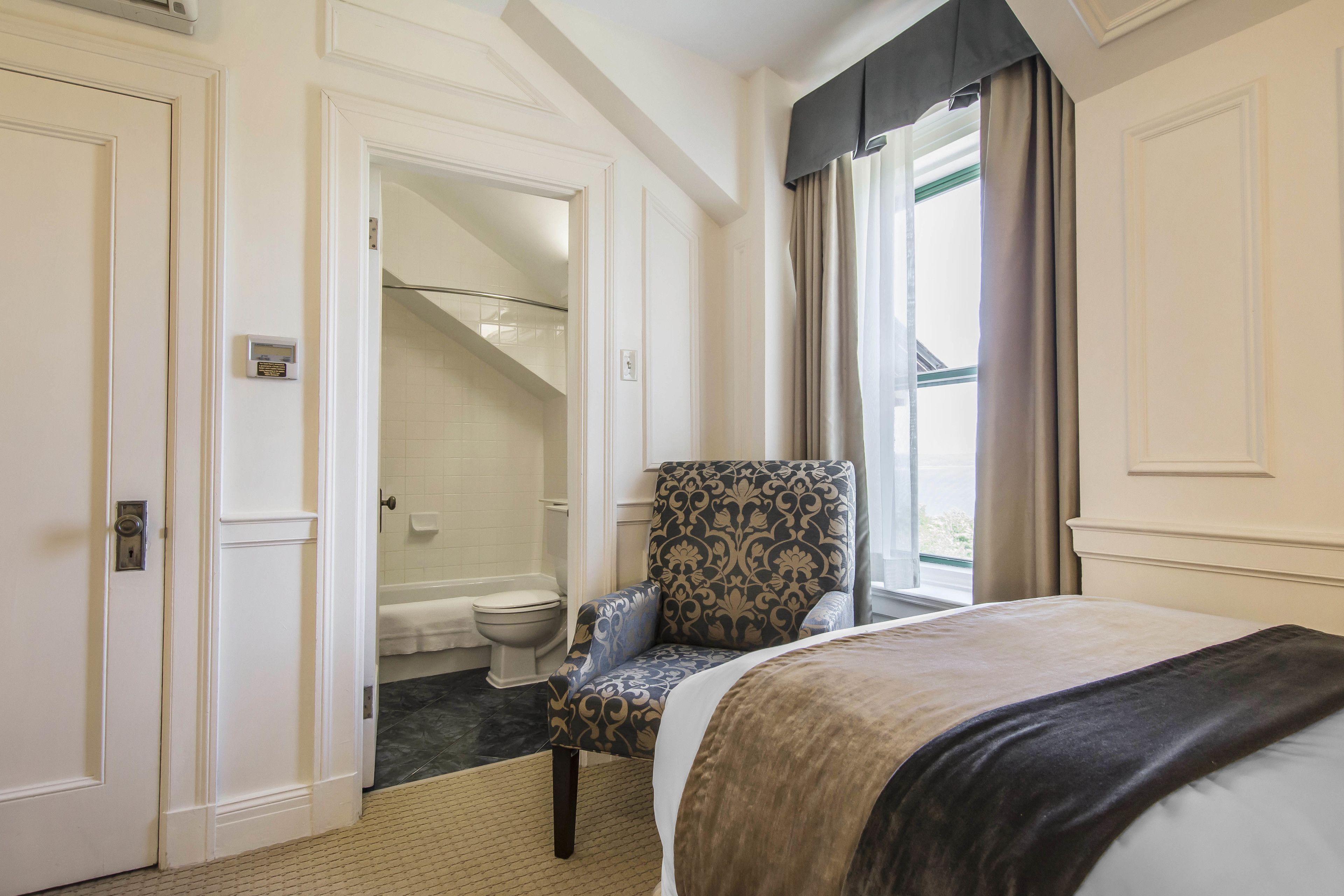 A hotel room with a bed, a chair, a bathroom with a toilet and shower, and a window with curtains.