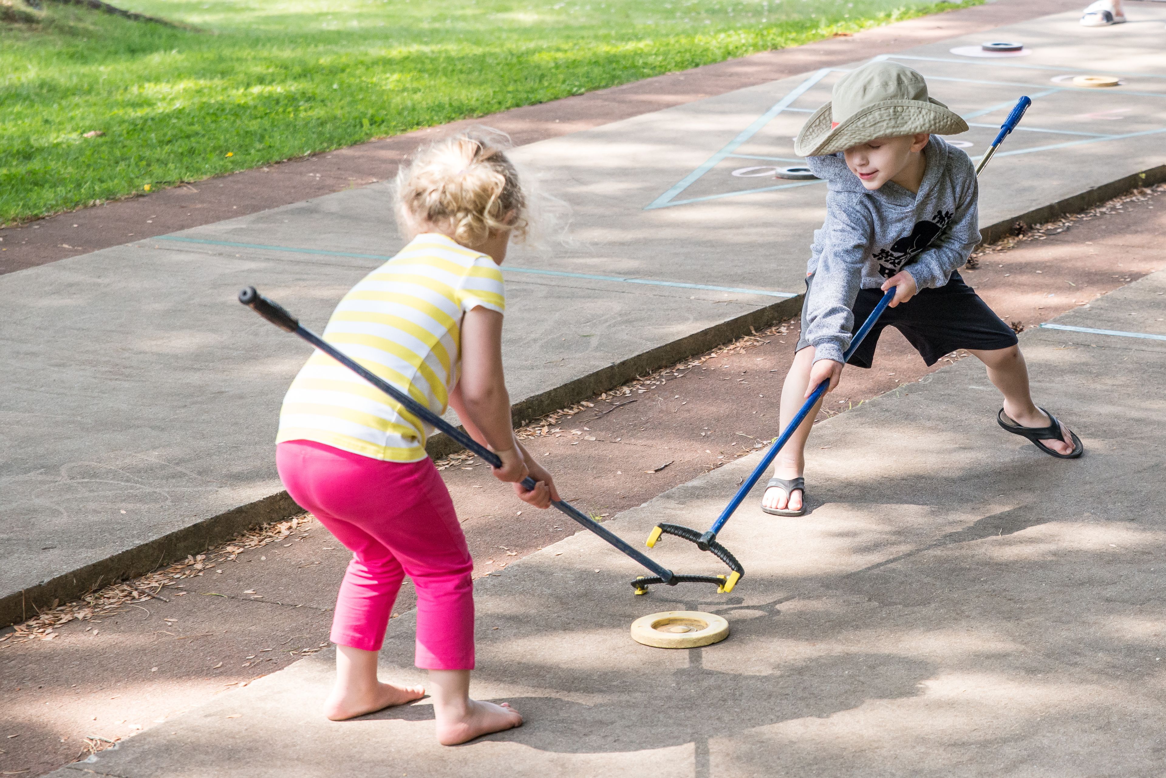 Two children playing shuffleboard on a concrete surface with a grassy area in the background.