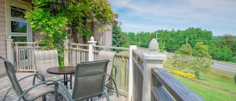 A wooden deck with outdoor furniture, a white railing, and a view of a road and greenery.