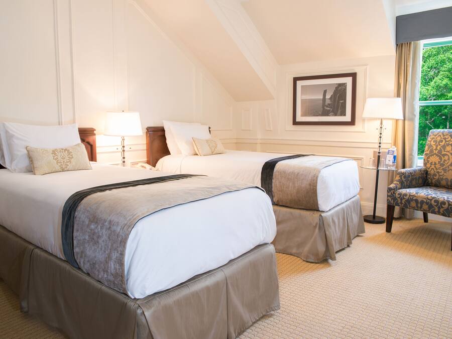 Une chambre d’hôtel avec deux lits, une chaise, un tableau encadré au mur et une fenêtre donnant sur un paysage verdoyant.