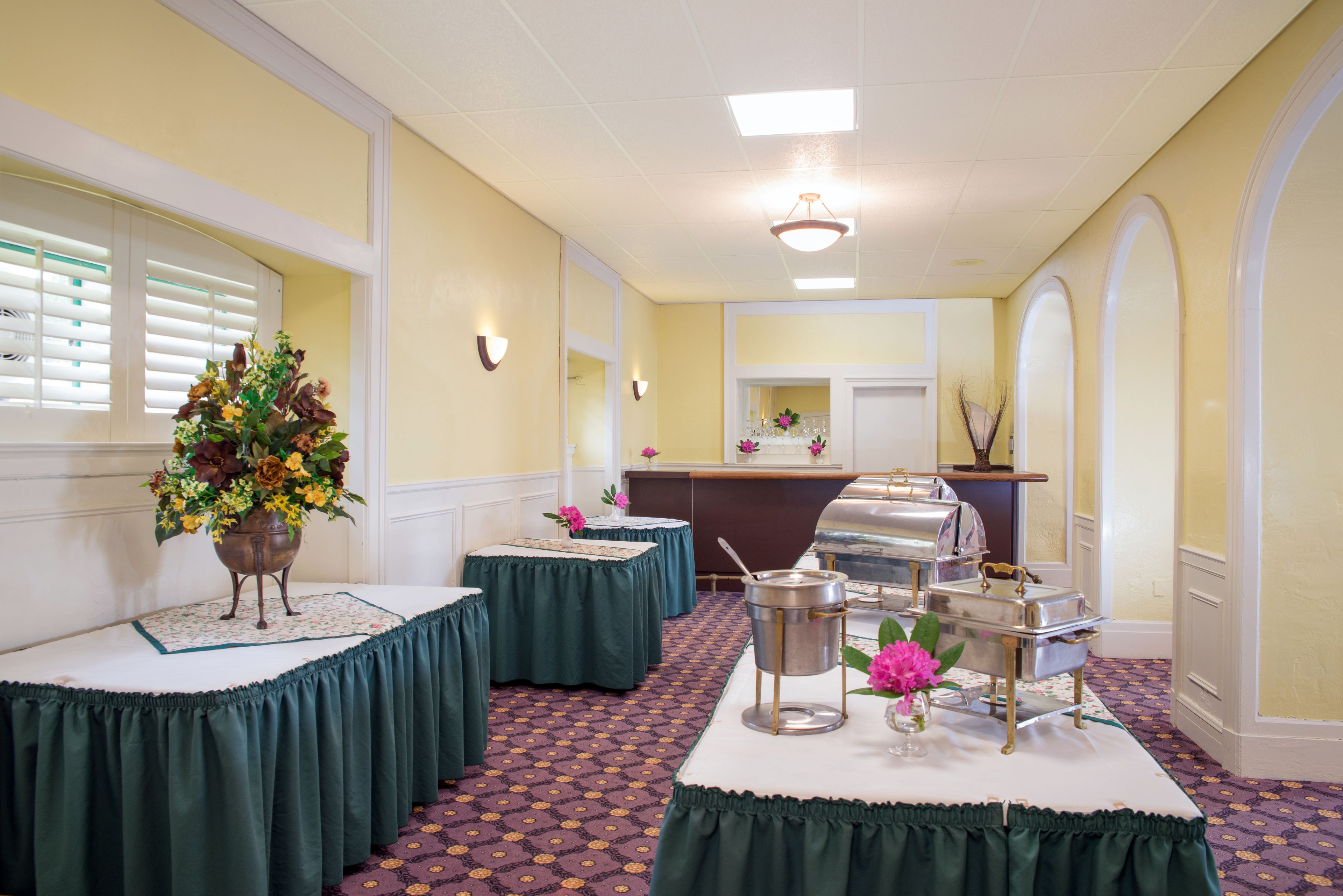 Une salle avec des tables recouvertes de nappes vertes, un buffet garni de plats de service en argent et un grand vase rempli de fleurs posé sur l’une des tables.