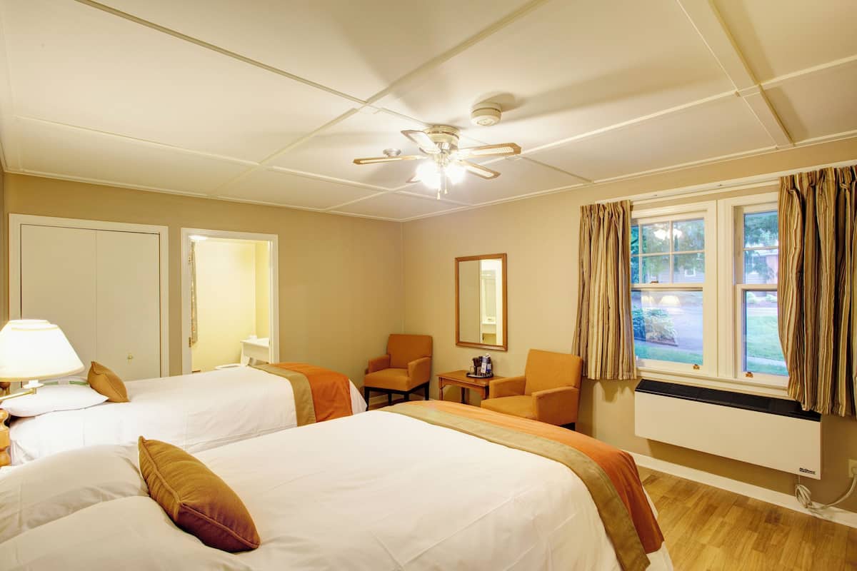A hotel room with two beds, a chair, a small table, a ceiling fan, and a window with curtains.