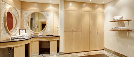 A modern bathroom with a curved vanity, large mirrors, a wooden wardrobe, and a bench.