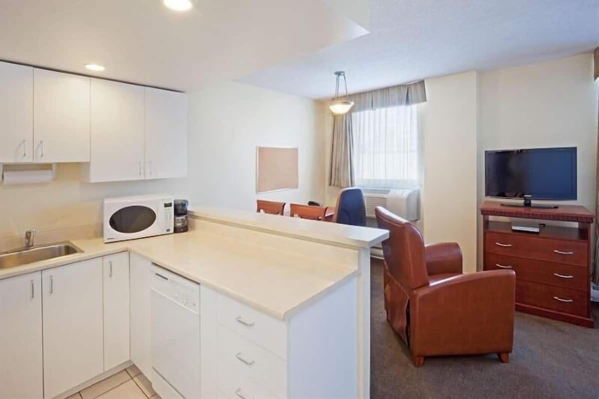 A compact hotel room with a kitchenette, a dining area, a TV, and a window with curtains.