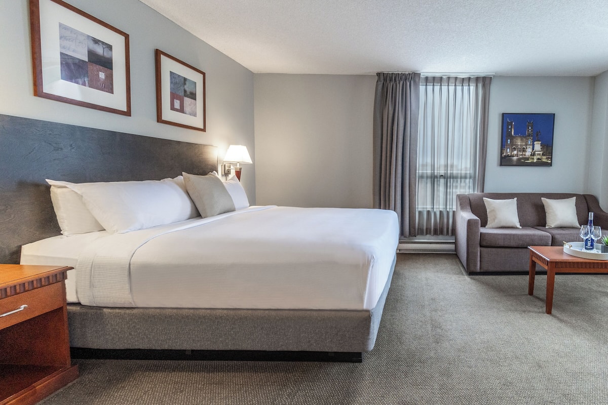 A hotel room with a large bed, a sofa, a small table with a glass, and framed pictures on the wall.
