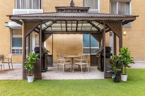 Une terrasse couverte équipée d’un barbecue, d’une table et de chaises, entourée de plantes en pot et d’un mur en briques.