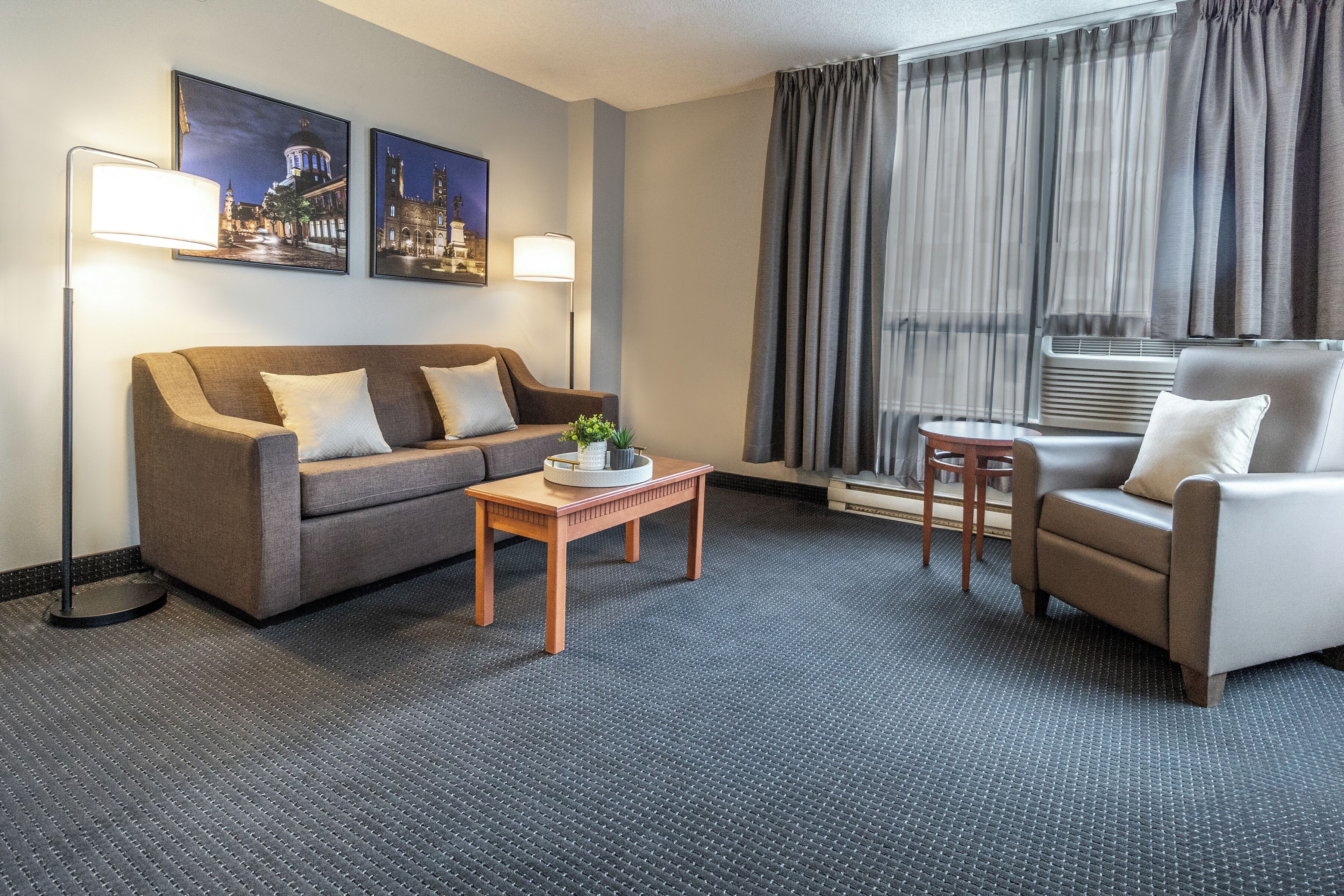A hotel room with a sofa, armchair, small table, floor lamp, and two framed pictures on the wall.