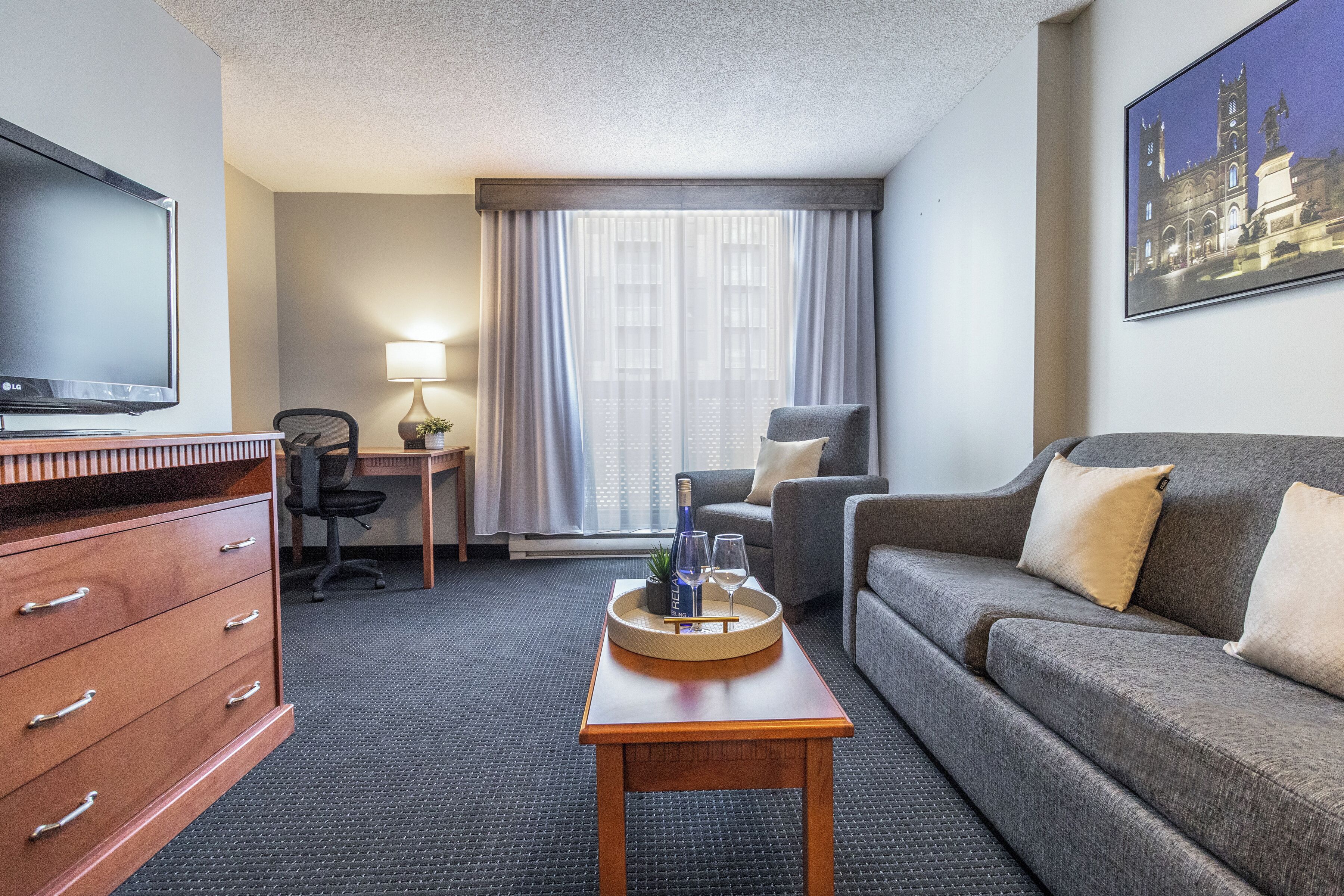 A hotel room with a flat-screen TV, a wooden dresser, a grey sofa, a small table with a tray, a chair, a desk with a lamp, and a large window with curtains.