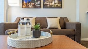 Hall d'hotel con un tavolo rotondo che espone due bottiglie di Hotel Sant Laurent Montreal, una piccola pianta in vaso e un vassoio decorativo. Sullo sfondo, un divano marrone e due quadri con scorci urbani.