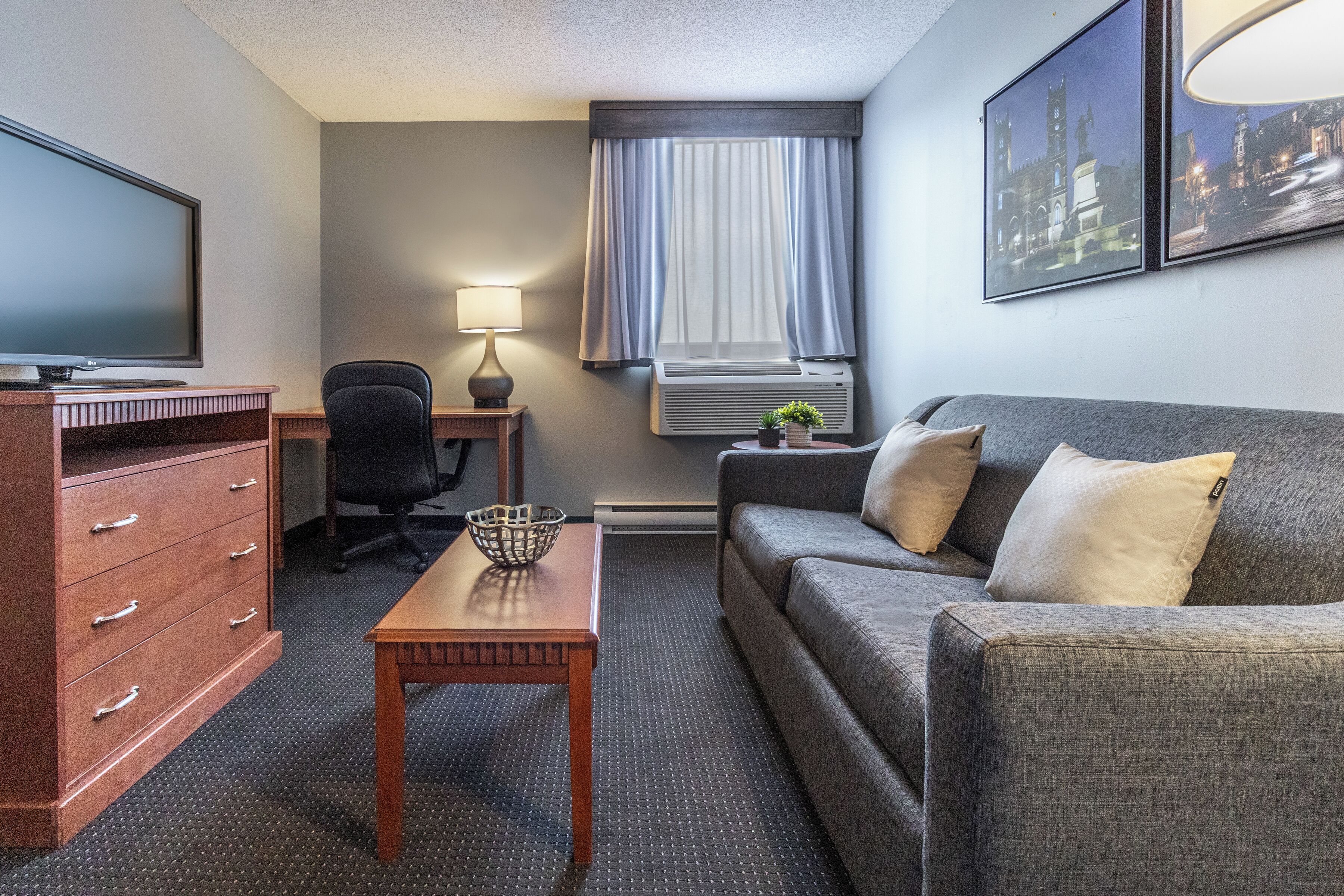 A hotel room with a television, desk, chair, sofa, and a window with curtains.