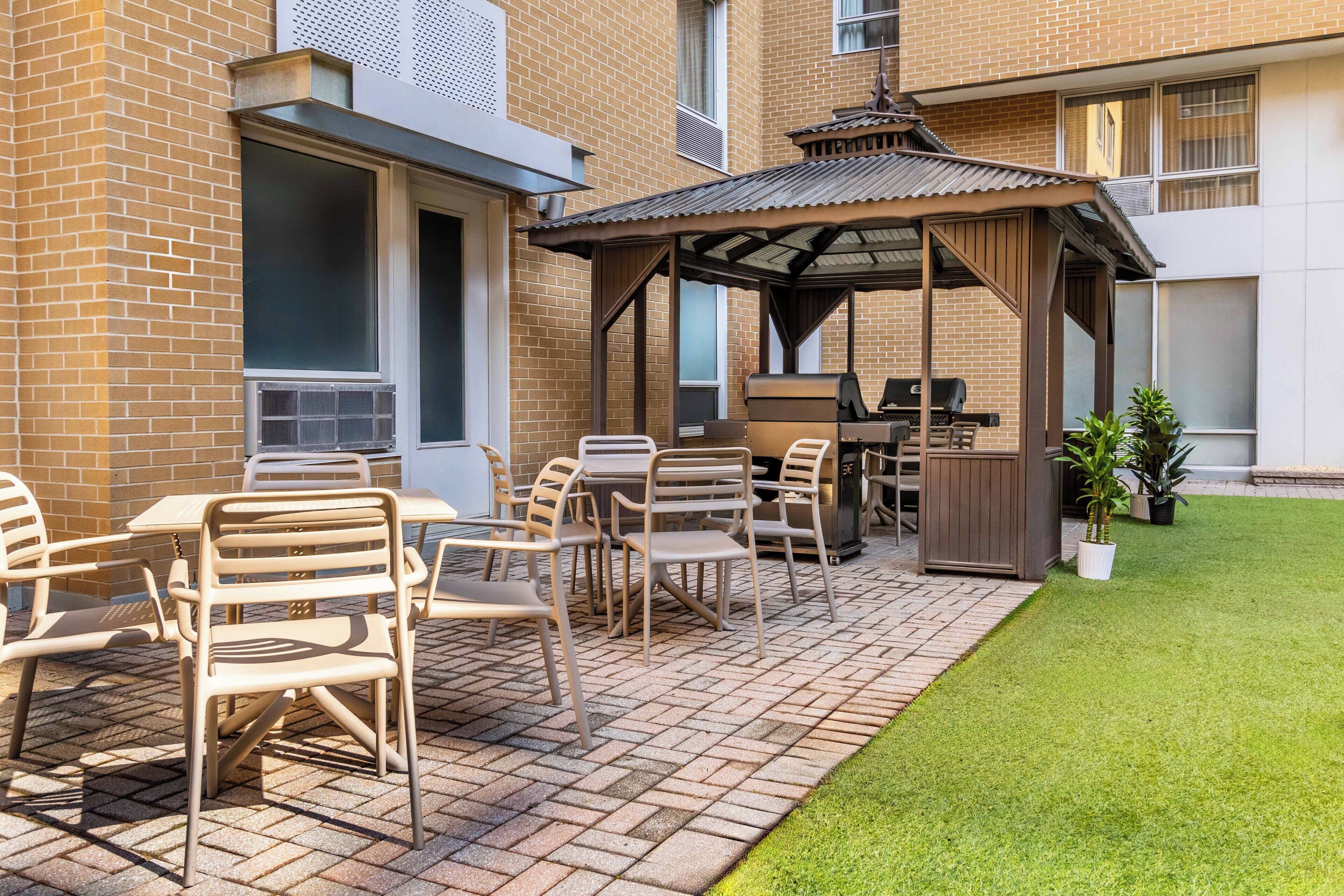 Un espace extérieur aménagé en terrasse, avec un sol en briques, des chaises en métal, un gazebo en bois et un barbecue.