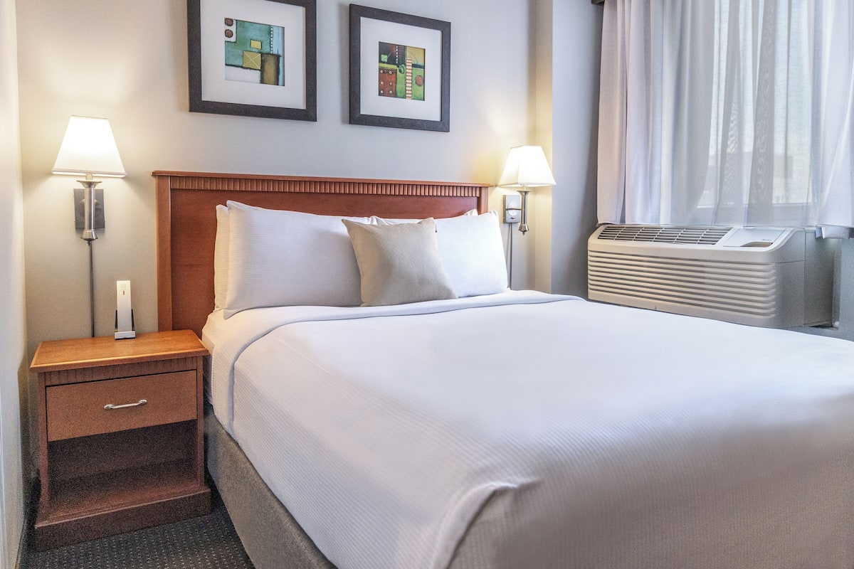 A neatly made hotel room with a large bed, two bedside lamps, a nightstand with a drawer, and an air conditioning unit.