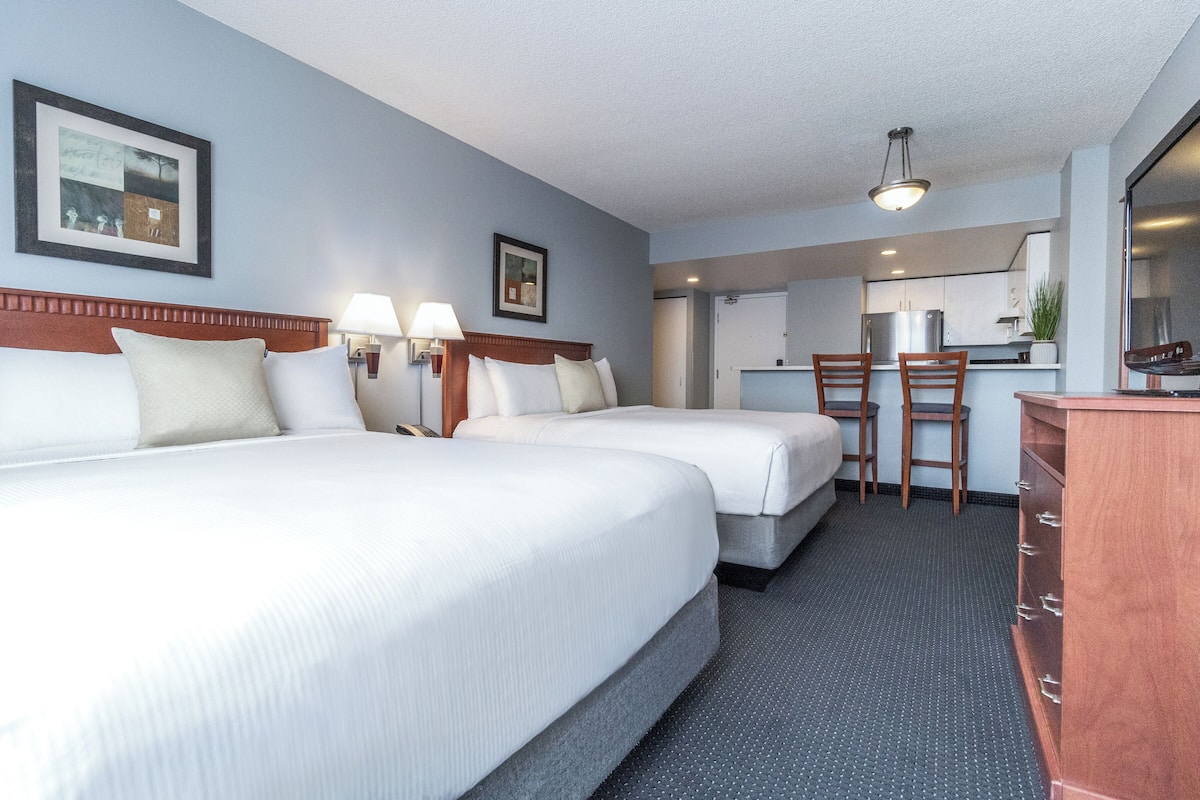 A hotel room with two beds, a desk, chairs, a dresser, and a kitchenette.