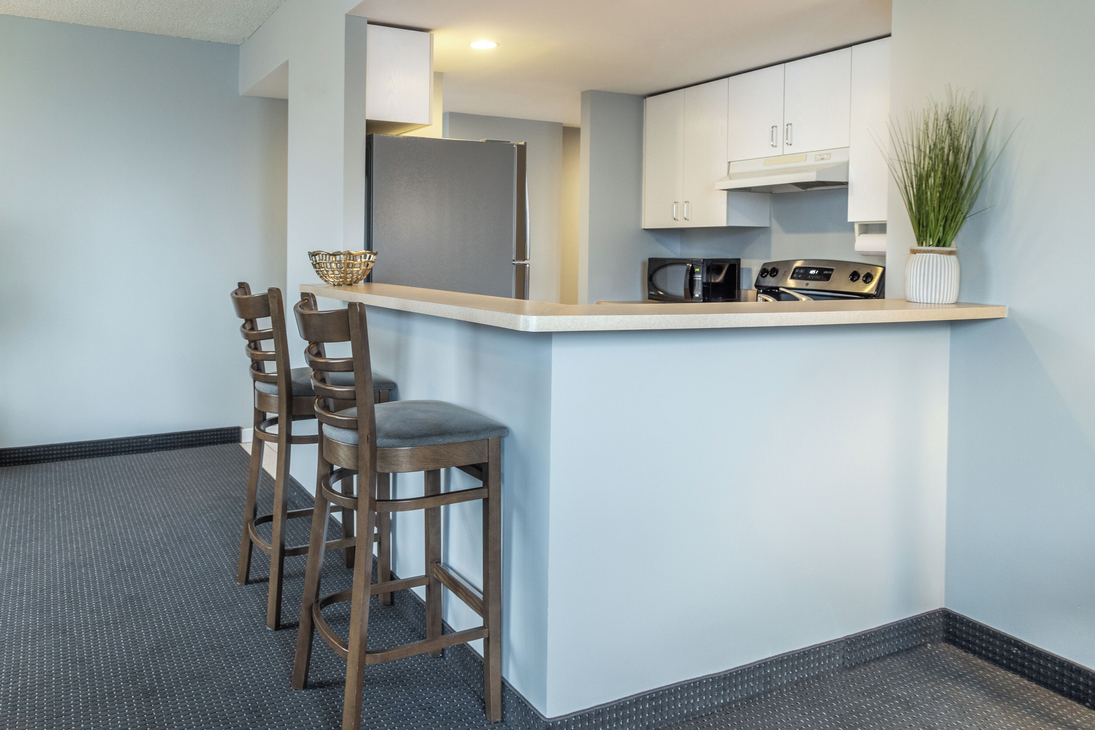 A compact kitchen with a bar area, equipped with a refrigerator, microwave, and stove. There are two wooden bar stools and a small potted plant on the counter.