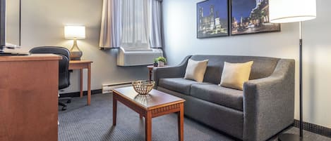 Une chambre d’hôtel avec un canapé gris, une table en bois sur laquelle se trouve un panier, un bureau avec une lampe et une grande fenêtre avec des rideaux.