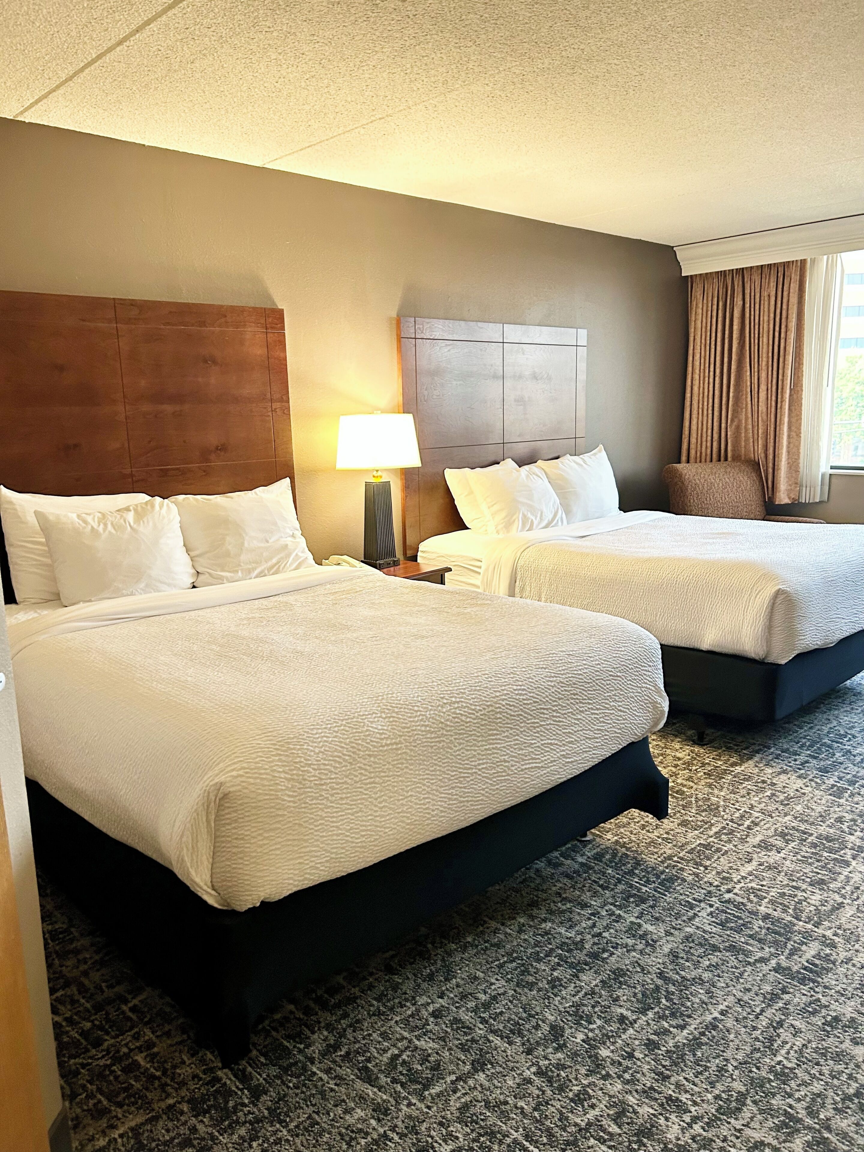 A hotel room with two beds, a nightstand with a lamp, a chair, and a view of the outdoors.