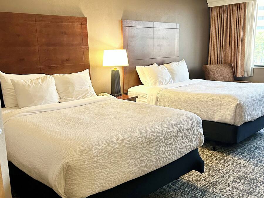 A hotel room with two beds, a nightstand with a lamp, a chair, and a view of the outdoors.