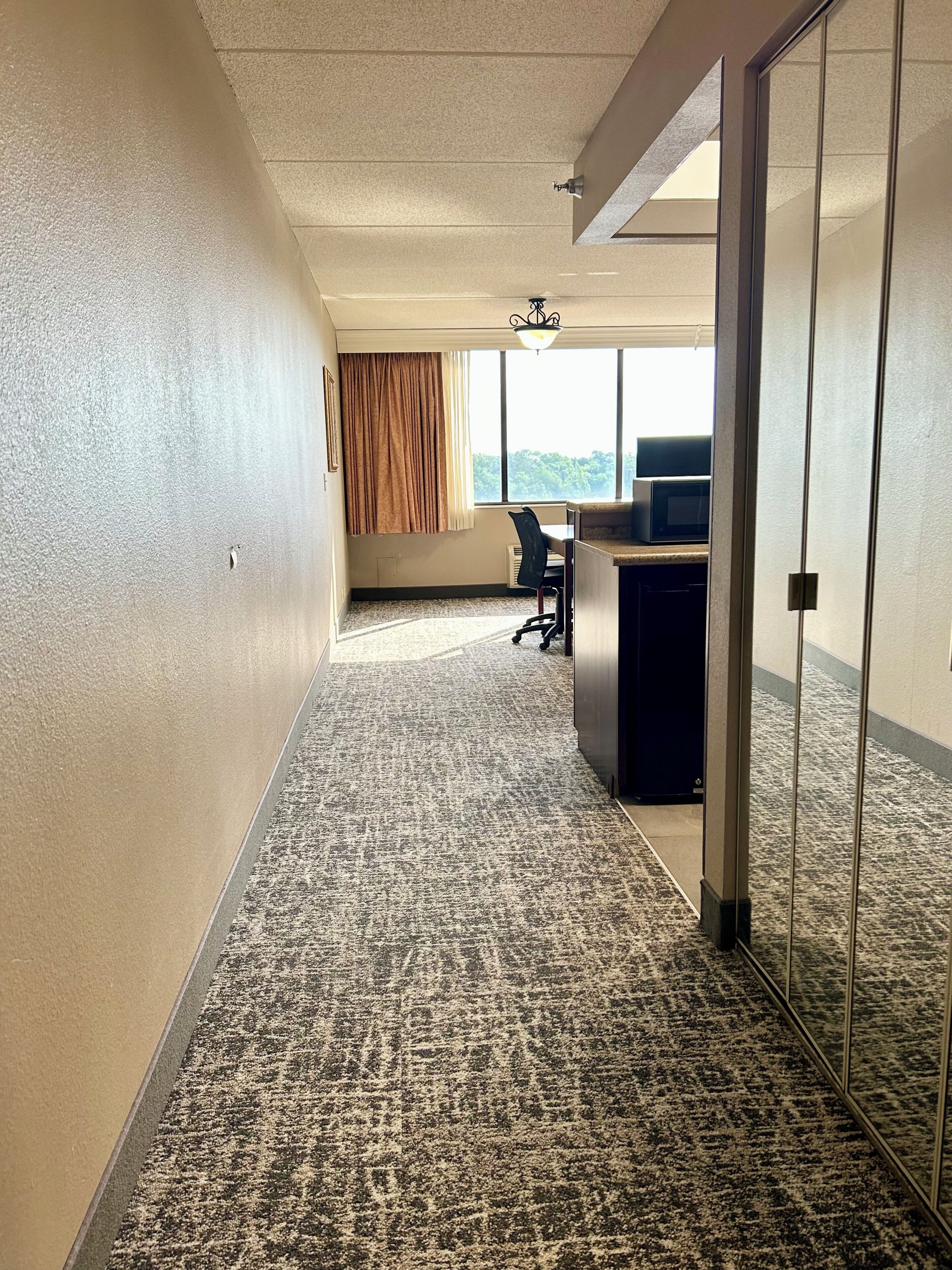 A hotel corridor with carpeted flooring, a desk with a computer, a chair, and a window with curtains.