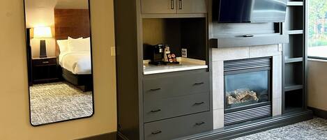 A hotel room with a large mirror, a wardrobe with drawers, a flat-screen TV, a fireplace, and a window with a view.