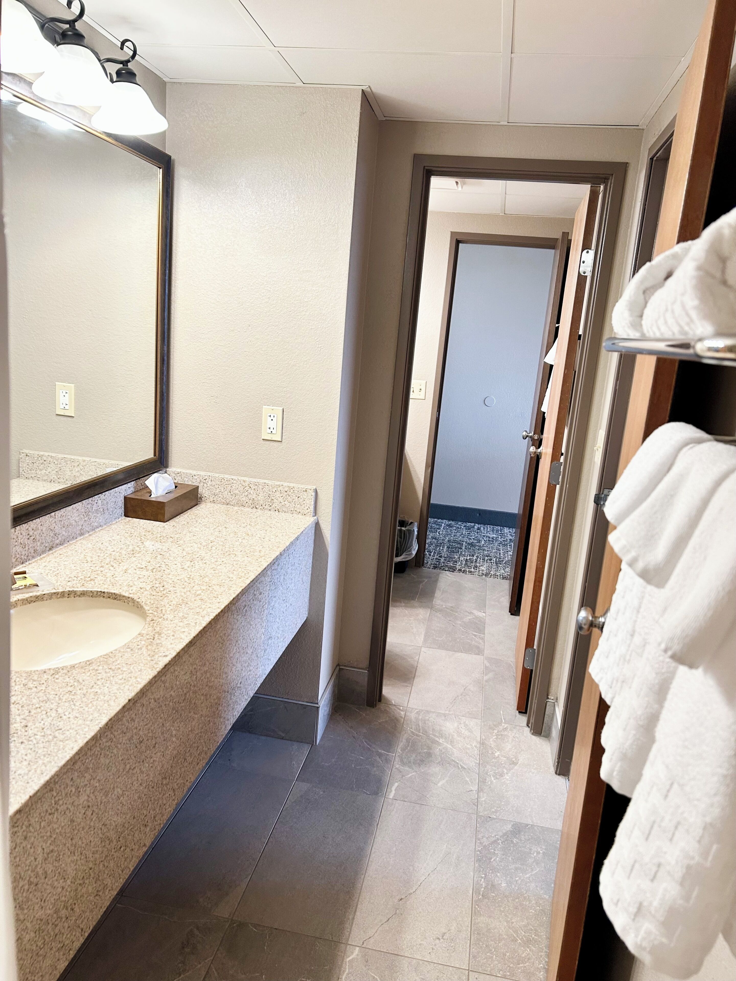 Un baño con lavamanos, espejo, perchero y una puerta que da a otra habitación.