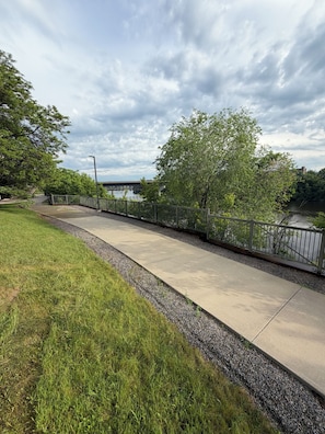 A concrete pathway alongside a river, with a metal fence and trees on one side.