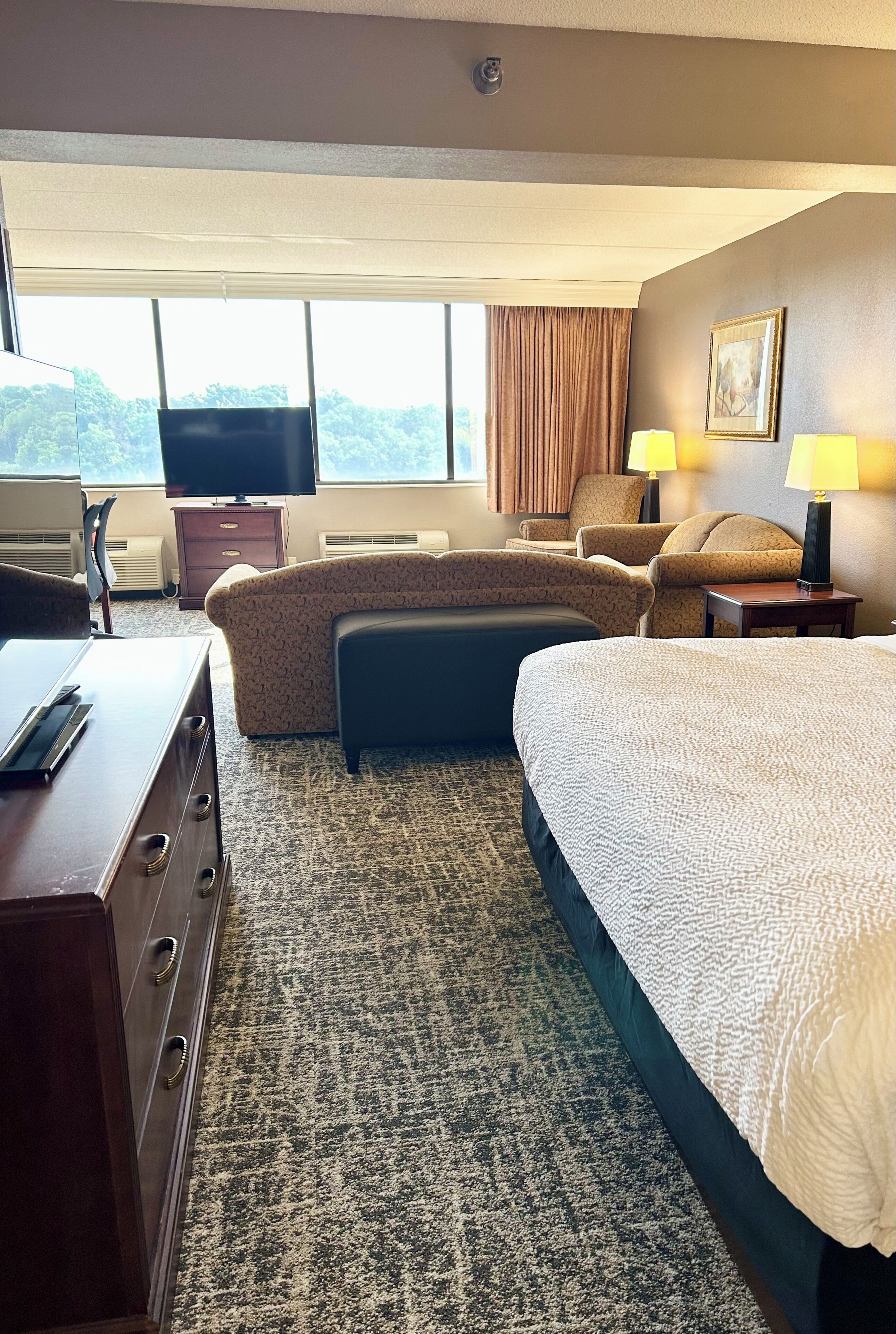Une chambre d’hôtel comprenant un lit, deux fauteuils, un bureau, une télévision et une fenêtre avec des rideaux.